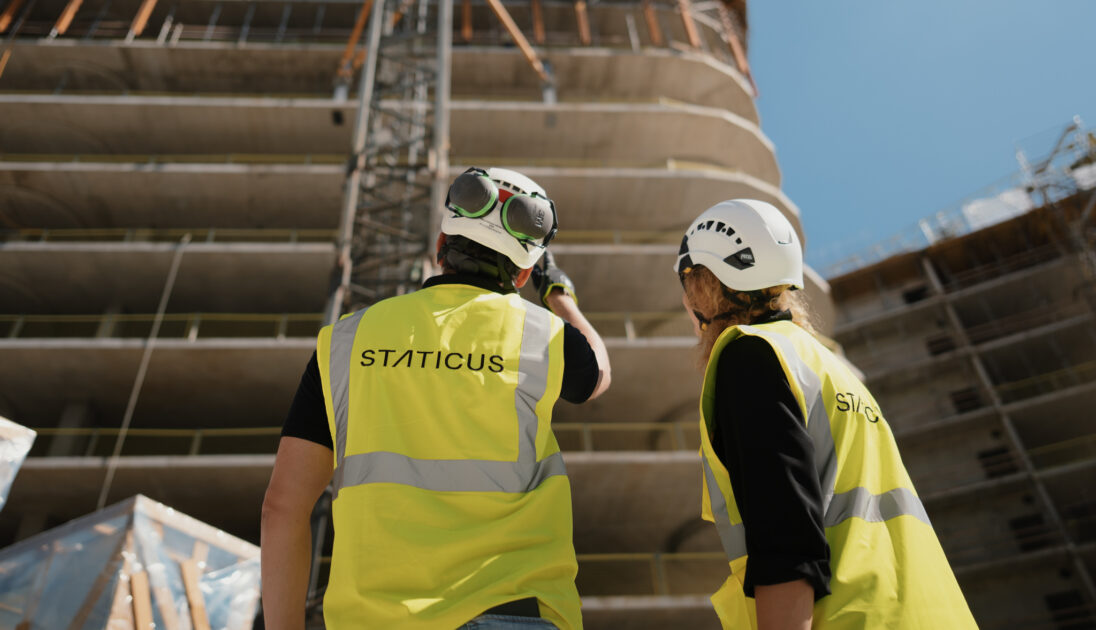 Two construction workers wearing high-visibility vests with "STATICUS" printed on the back, hard hats, and protective goggles are standing at a construction site. They are looking up at a large building under construction, with one worker pointing towards the structure, likely discussing the ongoing work.