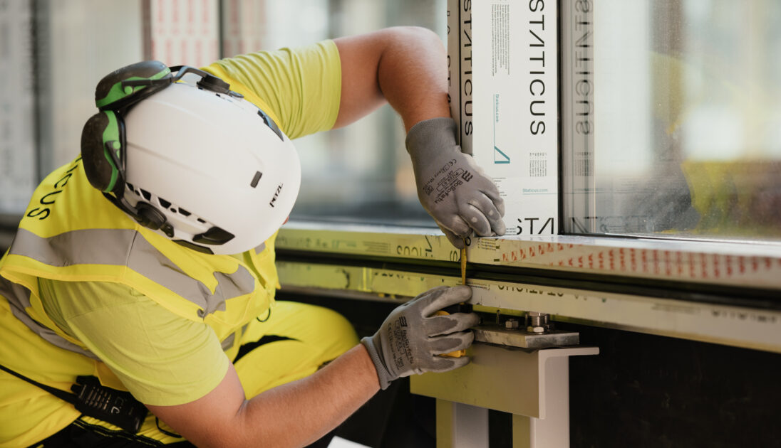 construction worker in a high-visibility vest, hard hat, and gloves is using a screwdriver to adjust or secure part of a window frame. The window is wrapped in material with "STATICUS" branding, indicating the installation process at the construction site.